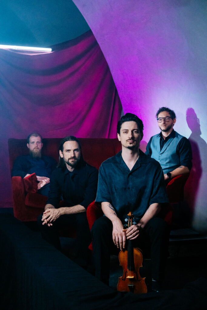 Die vier Bandmitglieder Jan Friedrich, Benedikt ter Braak, Tobias Paulus und Marco T. Alleata sitzen auf roten Samtsesseln vor einem überwiegend pinkfarbenen Hintergrund.